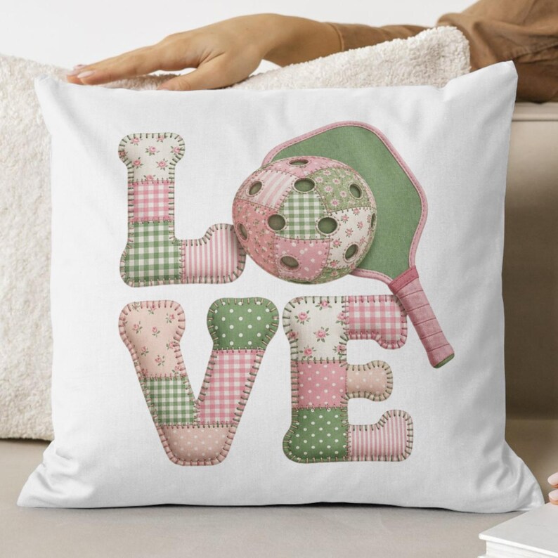 May include: White decorative pillow with the word "LOVE" in patchwork letters. The "O" is a pickleball, and a green and pink pickleball paddle is next to it. The letters and pickleball are made of pink and green floral and gingham fabrics.
