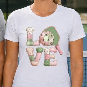 May include: White t-shirt featuring the word "LOVE" in patchwork style, with a pickleball and paddle design. The letters and pickleball are in pink, green, and white floral and gingham patterns. The paddle is green with a pink handle.