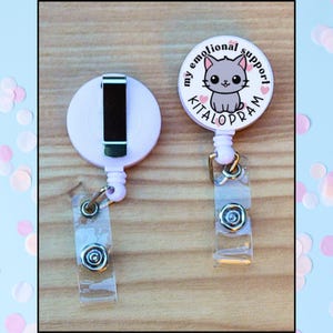 May include: Two light pink retractable badge holders. One has a black clip and the other has a badge with a gray cat illustration and the words "my emotional support KITALOPRAM". Both have clear plastic straps.