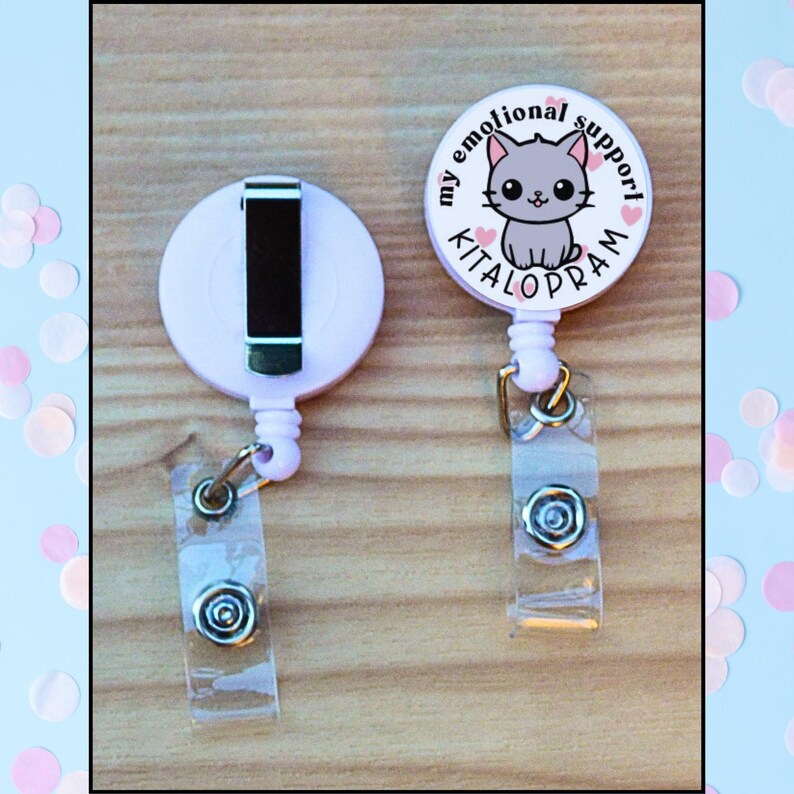 May include: Two light pink retractable badge holders. One has a black clip and the other has a badge with a gray cat illustration and the words "my emotional support KITALOPRAM". Both have clear plastic straps.