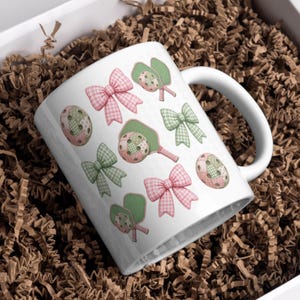 May include: White ceramic mug decorated with pink and green gingham bows, pickleball paddles, and balls. The design includes a mix of pink and green colors, creating a playful and sporty theme. The mug is set in a white box with brown paper filler.