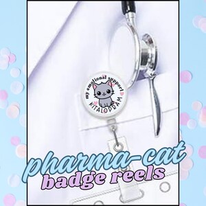 May include: A white badge reel with a cartoon cat design and the words "my emotional support" and "KITALOPRAM". The text "pharma-cat badge reels" is also displayed. A stethoscope is visible in the background.