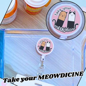 May include: A pink retractable badge reel with the text "take your meowdicine" and a cartoon illustration of two cats. One cat is orange and black, the other is white and black. The reel is attached to a clear plastic holder.