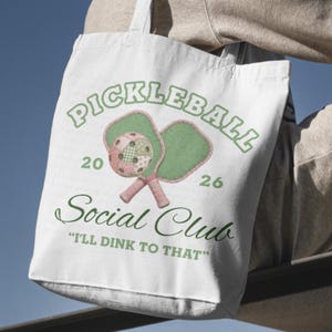 May include: White tote bag featuring a pickleball design. The design includes the word "PICKLEBALL" in green, along with two pickleball paddles and a ball. The text "Social Club" and the phrase "I'll dink to that" are also included.