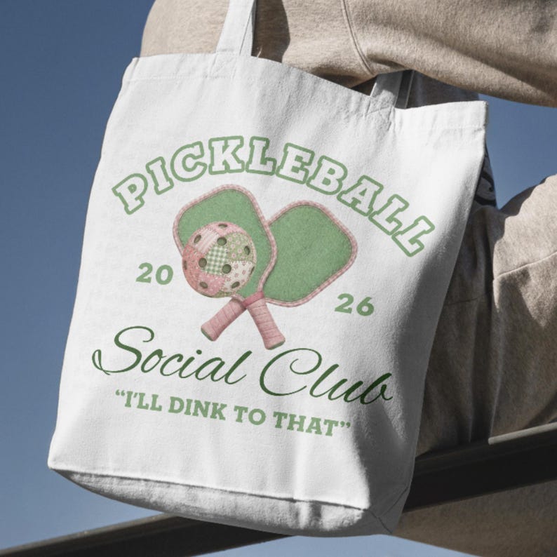 May include: White tote bag featuring a pickleball design. The design includes the word "PICKLEBALL" in green, along with two pickleball paddles and a ball. The text "Social Club" and the phrase "I'll dink to that" are also included.