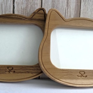 May include: Two wooden cat-shaped photo frames. The frames are light brown with a rectangular aperture for a photo and a white background. The cat's ears are at the top, and the frames have a carved nose and whiskers.