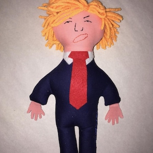 May include: A handmade doll with a felt face and yarn hair. The doll wears a navy blue suit with a red tie and has pink felt hands. The doll's face has a drawn expression.