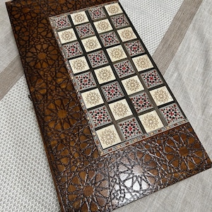 May include: A wooden backgammon board with a detailed geometric star pattern. The playing surface features a decorative inlaid design with squares of cream, black, red, and gold. The board is closed, showing the intricate exterior design.