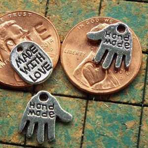 May include: Three silver charm tags with different sayings. One tag is oval shaped and says "Made with Love". The other two tags are shaped like hands and say "Hand Made".