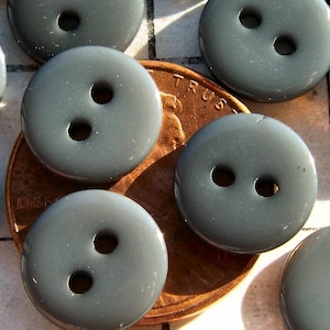 May include: A collection of gray, round buttons with two holes each. The buttons are arranged on a white surface, with a copper penny visible. The buttons are a solid, matte gray color.