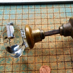 May include: A clear, faceted glass door knob set featuring two octagonal glass knobs connected by a central metal spindle. Each knob is attached to a tarnished brass-colored circular base. A copper penny is visible for scale reference.