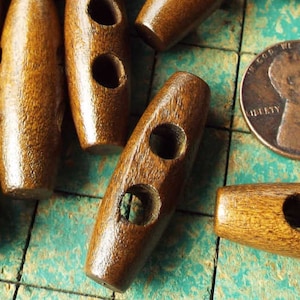May include: A close-up of a group of brown wooden toggle buttons. The buttons have two holes and are scattered on a green and white checkered surface. A copper penny is also visible in the image.