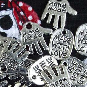 May include: A collection of silver-toned metal charms with various hand and heart shapes. The charms feature the text "Made with Love", "Hand Made", and "Appdw Pudh".