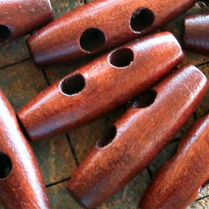 May include: A group of 12 brown wooden toggle buttons with two holes each. The buttons are arranged in a pile on a checkered surface.