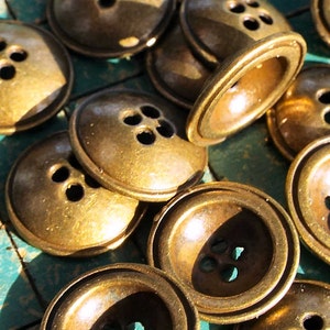 May include: A collection of 14 gold-toned metal buttons with four holes each. The buttons are arranged in a random pattern on a green and white checkered surface.