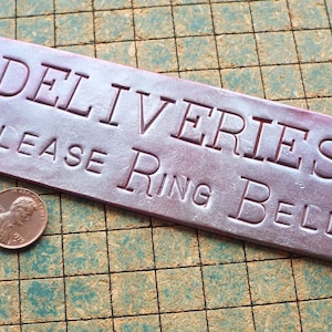 May include: A silver metal sign with rounded corners and holes for hanging. The sign reads "Deliveries Please Ring Bell" in a raised, embossed font.