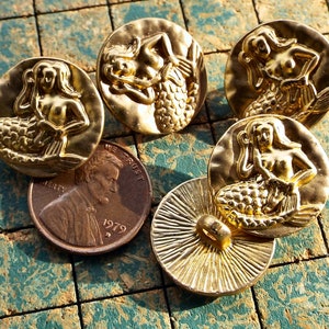 May include: Five gold-toned metal buttons featuring a mermaid design. Each button has a raised, detailed mermaid figure with a scaly tail and flowing hair.