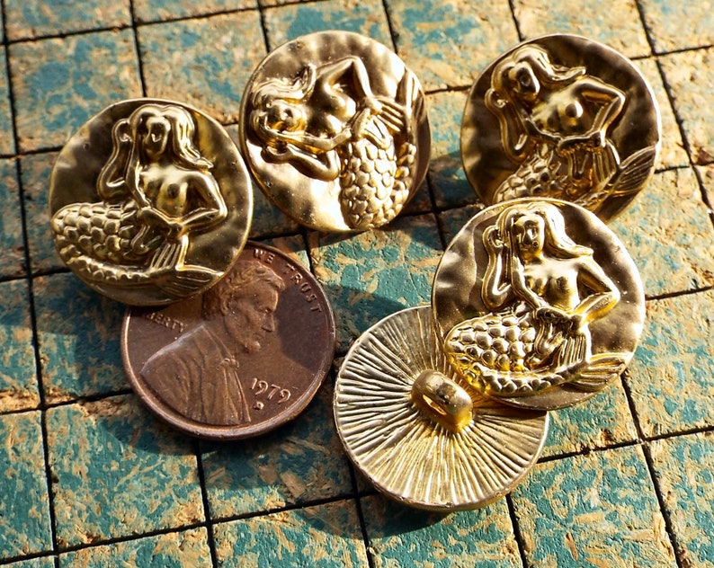May include: Five gold-toned metal buttons featuring a mermaid design. Each button has a raised, detailed mermaid figure with a scaly tail and flowing hair.