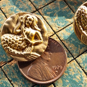 May include: Two gold-tone mermaid buttons with a raised design. The mermaid is depicted with long flowing hair and a tail. The buttons are approximately 1 inch in diameter.