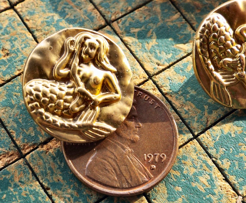 May include: Two gold-tone mermaid buttons with a raised design. The mermaid is depicted with long flowing hair and a tail. The buttons are approximately 1 inch in diameter.
