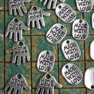 May include: A collection of silver metal charms with the words "Hand Made" and "Made With Love" engraved on them. The charms are arranged on a green and white checkered surface.