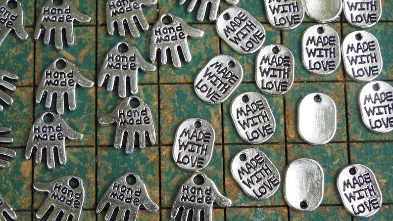 May include: A collection of silver metal charms with the words "Hand Made" and "Made With Love" engraved on them. The charms are arranged on a green and white checkered surface.