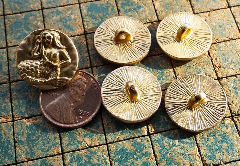 May include: Five gold-toned buttons with a mermaid design and a sunburst design. The mermaid button features a raised design of a mermaid with a tail and long hair. The sunburst buttons have a raised design of a sunburst with a loop at the top.