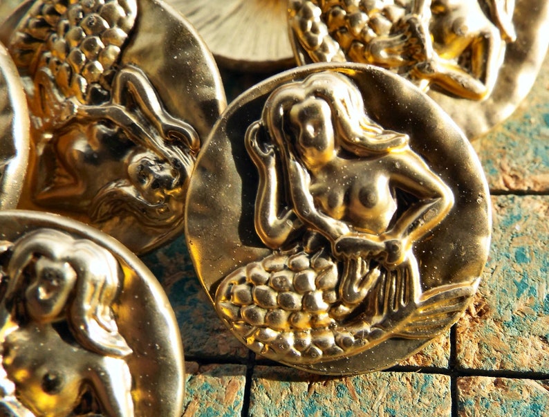 May include: A set of four gold-tone metal buttons featuring a mermaid design. Each button has a raised, detailed mermaid figure with a scaly tail and flowing hair.