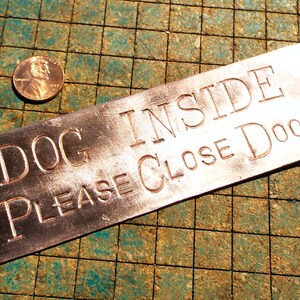 May include: A rectangular metal sign with the words "DOG INSIDE PLEASE CLOSE DOOR" engraved in capital letters. The sign has rounded corners and holes for mounting. A penny is next to the sign.