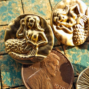 May include: Two gold-tone mermaid buttons with raised details. The mermaids are depicted with flowing hair and tails. The buttons are round and have a smooth, shiny finish.