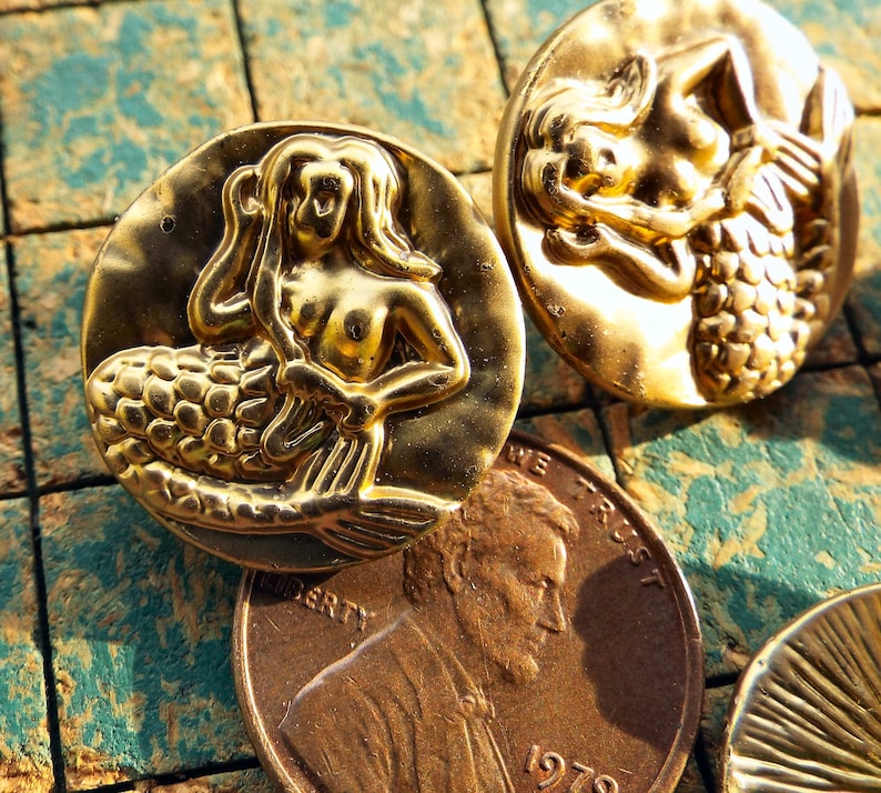 May include: Two gold-tone mermaid buttons with raised details. The mermaids are depicted with flowing hair and tails. The buttons are round and have a smooth, shiny finish.