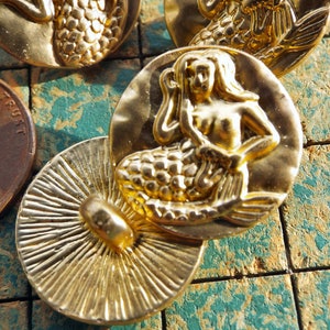 May include: Gold-tone metal mermaid button with a detailed design. The mermaid is depicted with long flowing hair and a scaly tail. The button has a smooth, shiny finish.