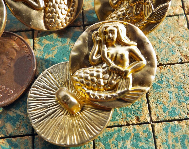 May include: Gold-tone metal mermaid button with a detailed design. The mermaid is depicted with long flowing hair and a scaly tail. The button has a smooth, shiny finish.