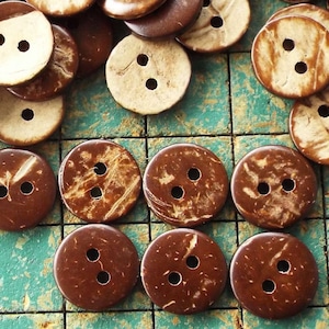 May include: A collection of brown and white coconut shell buttons with two holes each. The buttons are arranged in a grid pattern on a green and brown surface.
