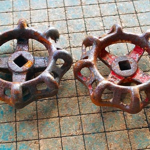 May include: Two antique, rusty metal valve handles. The handles have a circular design with a square center and are weathered with shades of brown and red. They rest on a blue and green grid surface.