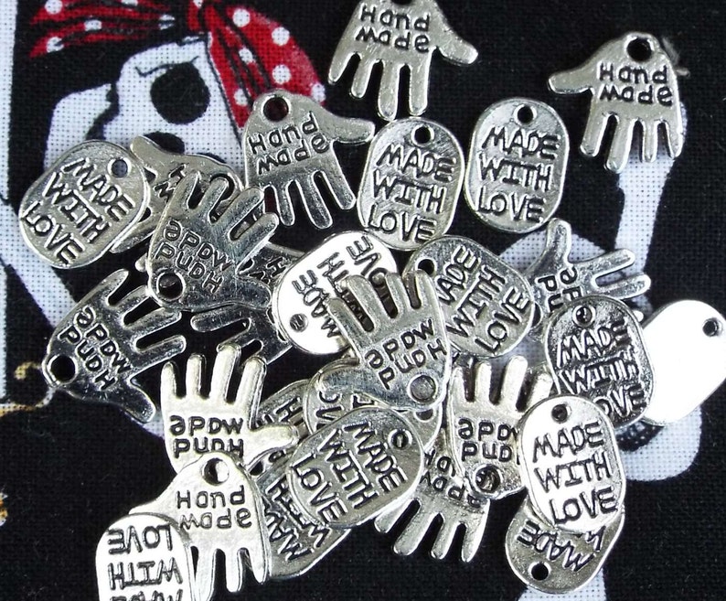 May include: A collection of silver-toned metal charms with various hand and heart shapes. The charms feature the text "Made with Love", "Hand Made", and "Appdw Pudh".