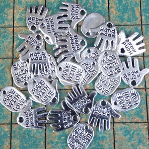May include: A collection of silver charm tags with various hand and oval shapes. The charms feature the text "Made with Love" and "Hand Made".