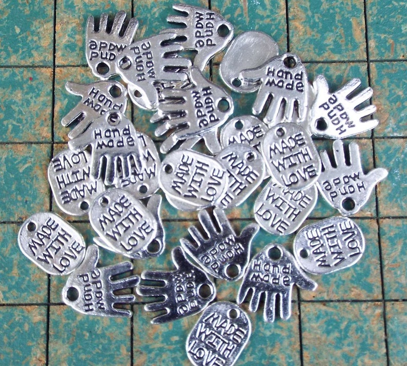 May include: A collection of silver charm tags with various hand and oval shapes. The charms feature the text "Made with Love" and "Hand Made".
