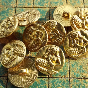 May include: Ten gold-tone metal buttons featuring a mermaid design. The buttons are round and have a raised design of a mermaid with a tail and flowing hair.