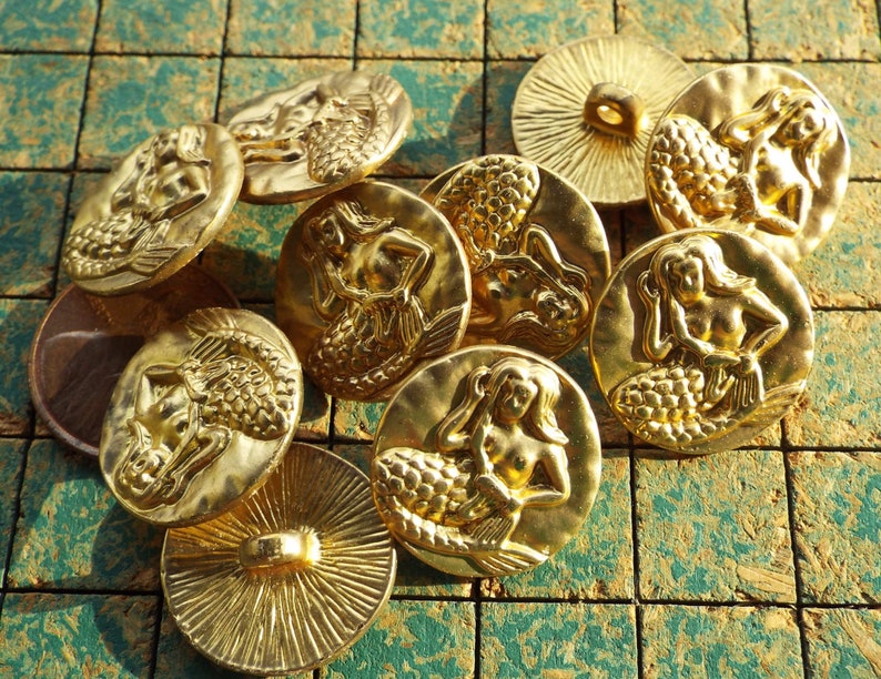 May include: Ten gold-tone metal buttons featuring a mermaid design. The buttons are round and have a raised design of a mermaid with a tail and flowing hair.