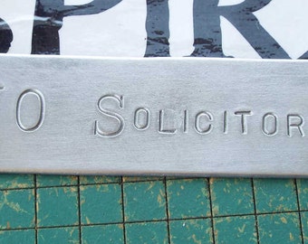 May include: A silver metal sign with the text "NO SOLICITORS" engraved on it.