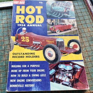 May include: A vintage "Hot Rod" magazine from 1954, featuring a red race car with the number 25. The cover has a blue and yellow background with the magazine title in large yellow letters. Additional images of cars and engines are also visible.