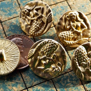May include: Five gold-toned metal buttons with a mermaid design. The mermaids are depicted in various poses, with their tails and hair flowing. The buttons are round and have a smooth, shiny finish.