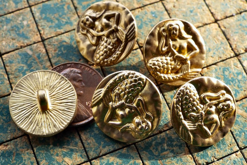 May include: Five gold-toned metal buttons with a mermaid design. The mermaids are depicted in various poses, with their tails and hair flowing. The buttons are round and have a smooth, shiny finish.