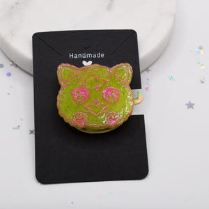 May include: A green and pink resin pin with a cartoon cat face. The pin says "Handmade" on the back.