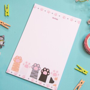 May include: A white notepad with a pink paw print border and paw print accents. The notepad has the word "Notes" printed at the top. Decorative items include a roll of pink patterned tape, miniature cat figurines, and clothespins.