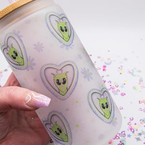 May include: A frosted glass tumbler with a bamboo lid and a straw. The tumbler features a repeating pattern of hearts with cartoon alien faces inside. The hearts are outlined in blue and have a purple background. The background of the tumbler is white with a light purple and white floral pattern.