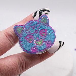 May include: A blue and pink cat-shaped phone grip with glitter accents. The grip features a cartoon cat face with green ears, pink eyes, and a heart-shaped nose. The edges are outlined in pink.