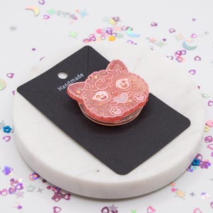 May include: Pink resin cat charm with glitter and a heart design. The charm is on a black card with the word "Handmade" written on it.