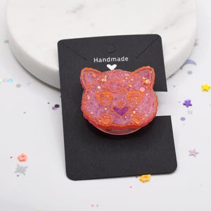 May include: A pink and orange resin cat charm with glitter embedded in the resin. The charm is on a black card with the word "Handmade" written on it.